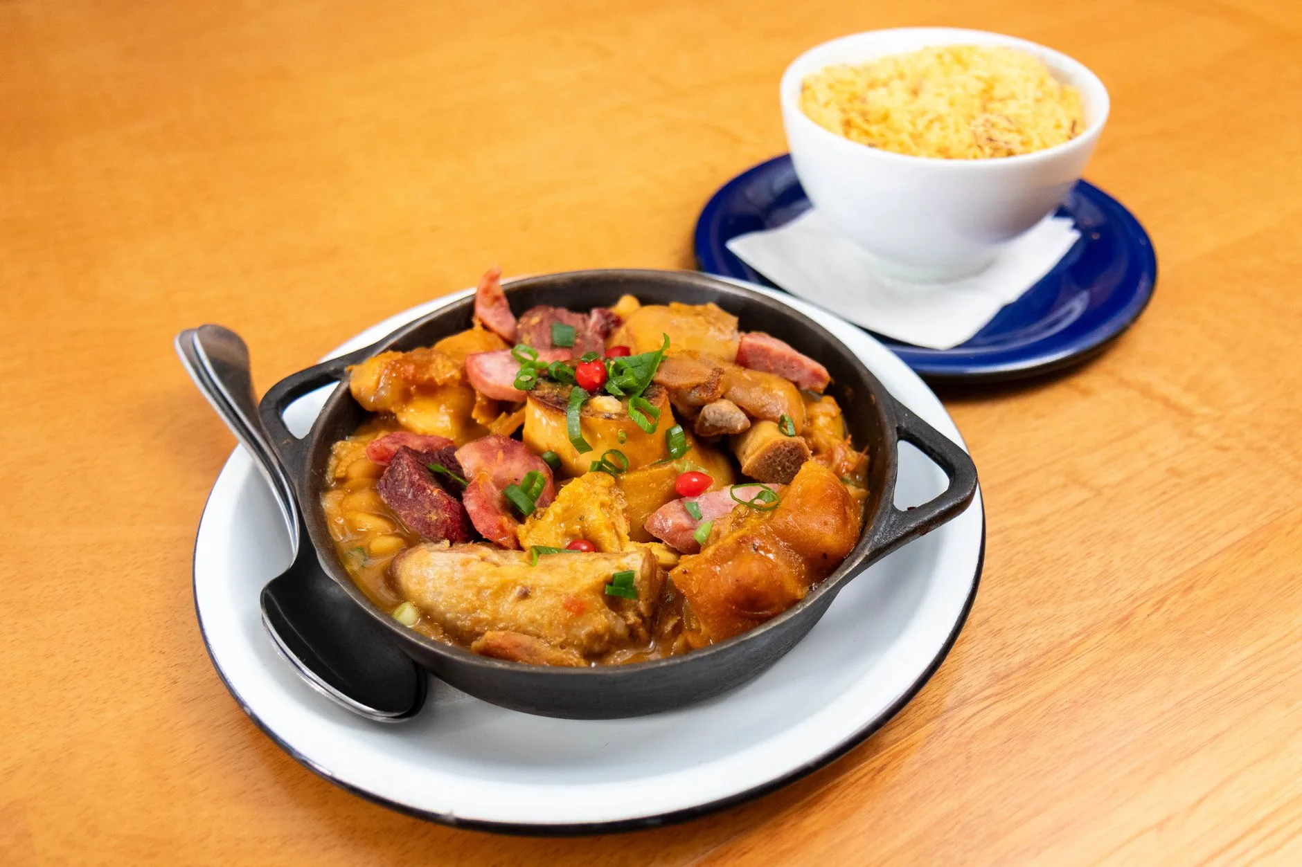 Feijoada brasileira servida em panela preta com arroz ao fundo, em uma mesa de restaurante