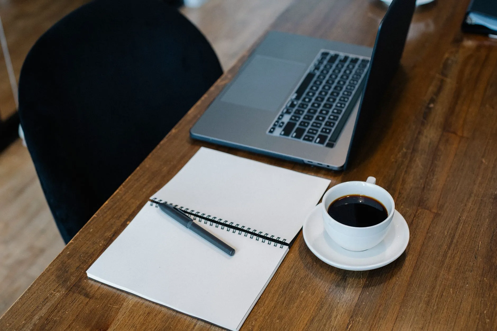 Notebook aberto ao lado de caderno em branco e xícara de café sobre balcão de madeira