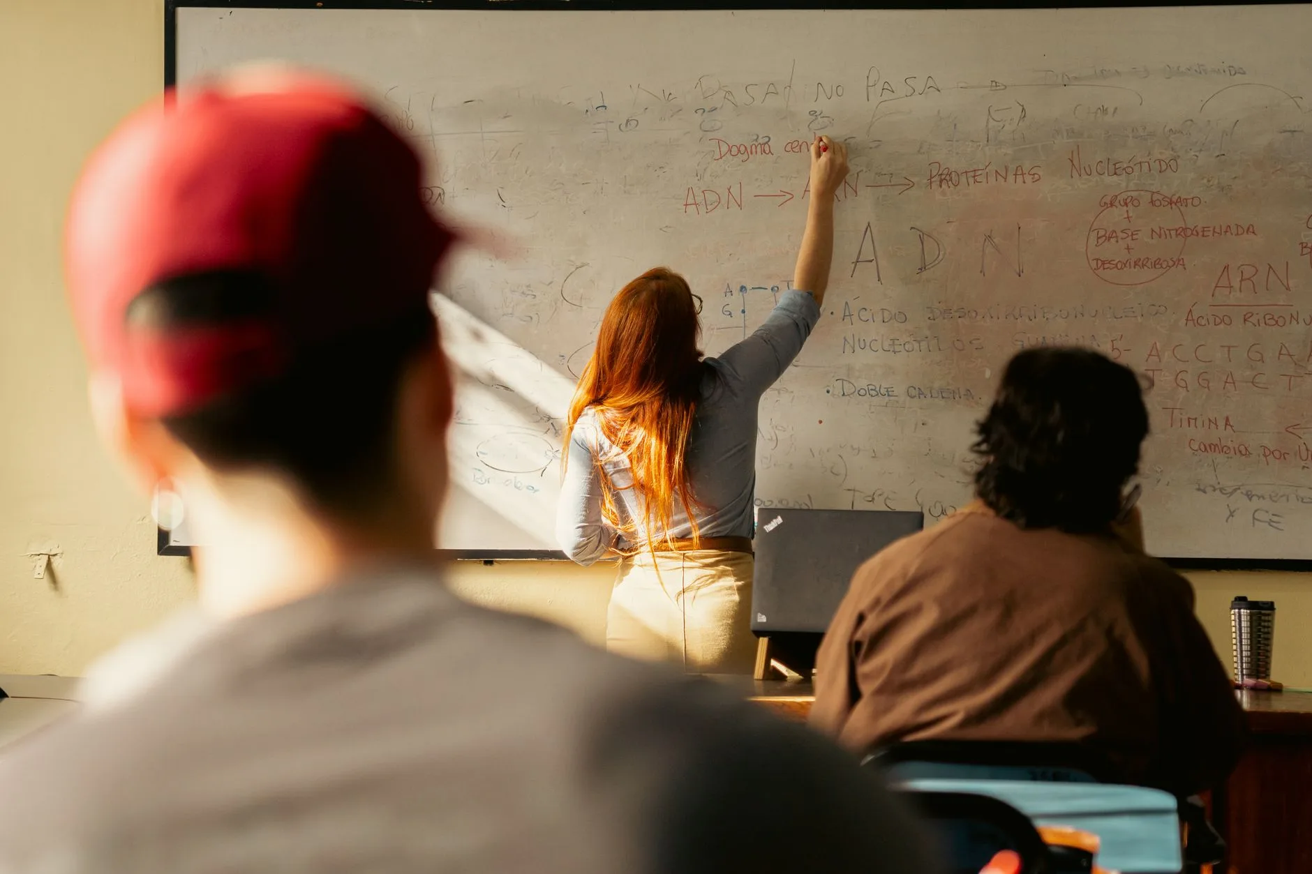 Professora escrevendo no quadro branco durante uma aula, com estudantes ao fundo.