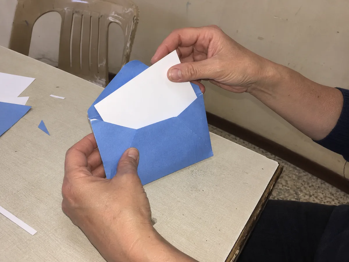 Mãos seguram um envelope azul aberto e inserem um cartão branco, com retalhos de papel sobre uma mesa em uma sala cotidiana.